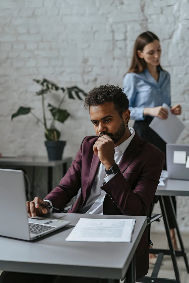 A business professional reviews a document at his laptop while a colleague stands behind him.